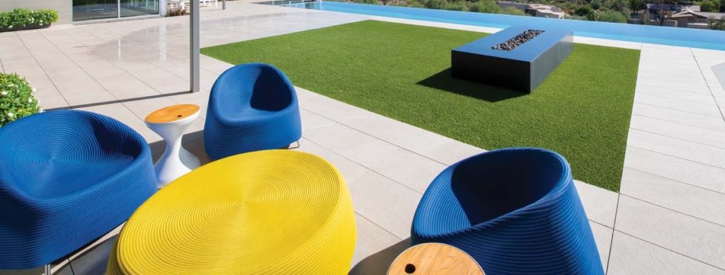 A vibrant outdoor seating area with bold blue chairs and a yellow table set on a wooden deck next to a green lawn and a pool The modern design and bright colors create a playful and inviting atmosphere