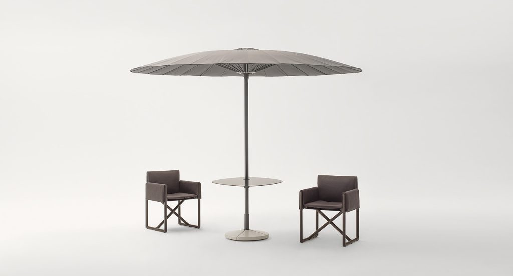 Beige flat shaped Bistro Umbrella with fabric shade and steel leg nexto to two chairs on a white background.