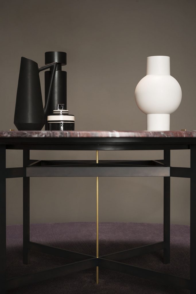 A Placè Dining Table. Base in brass with bronzing oxidation on the flat part and satin finish on the thickness, metal base varnished with burnished effect and satin brass details. Leg connection plate in matte black metal. Rocky mountain stone top with water-repellent protective oil with reinforcing aluminium honeycomb support. Decorative inserts in satin-finished brass with wax finish i na grey room.