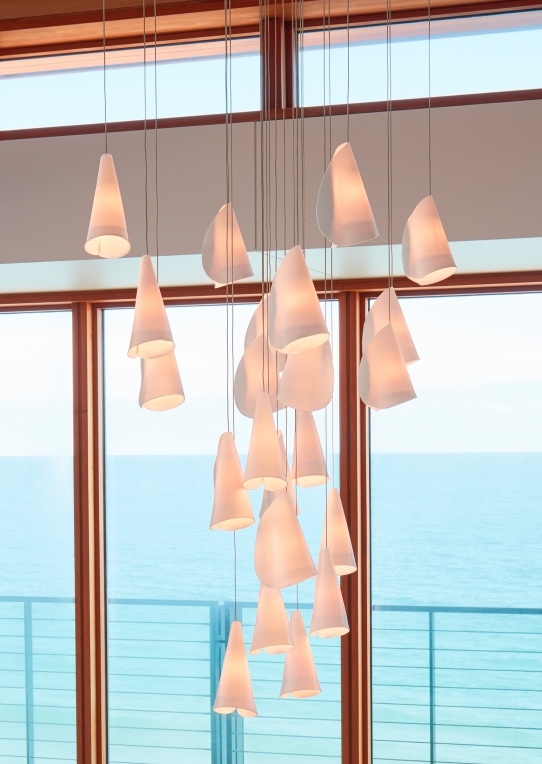 A Twenty One lamp made of porcelain, blown borosilicate glass, braided metal coaxial cable, electrical components, brushed white powder coated canopy in a room with the ocean background,