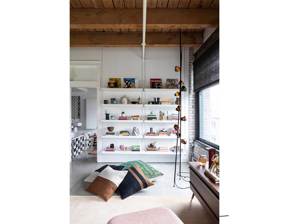Boc Stem Column Floor Light stands tall in a contemporary loft interior, its deep amber glass shades glowing warmly against a matte black stem. Positioned by a bookshelf and large window, it adds a sculptural accent to the bright, open space.