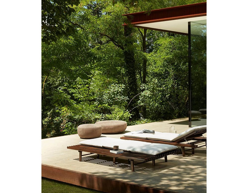 Two 10th Clove Outdoor Poufs are placed on a patio overlooking a lush green forest, alongside a coffee table and lounge chairs