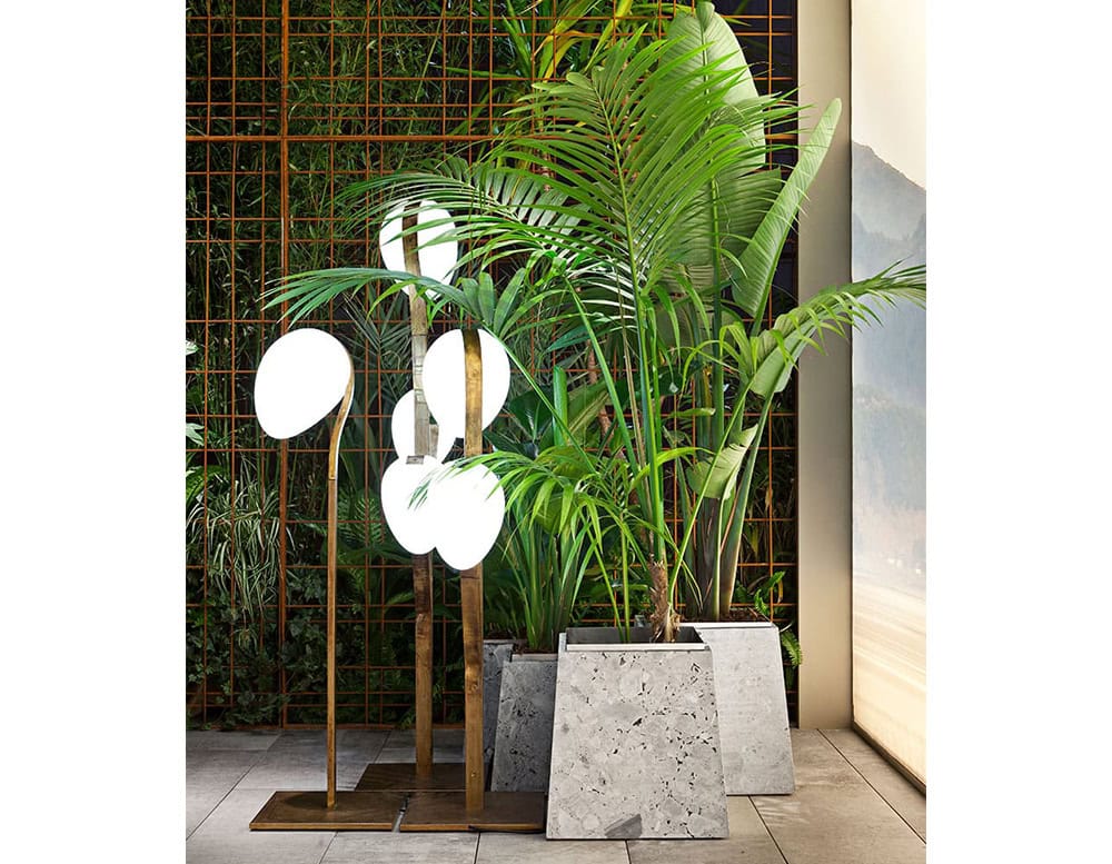A 10th Tree Light Outdoor Floor Light with multiple white, orb-shaped diffusers on slender stems is partially visible behind a potted plant in a room with a textured wall