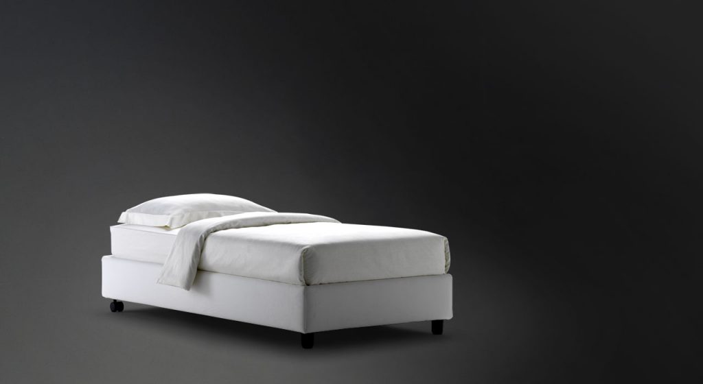 Sommier Single bed. White base with four black wheels and a mattress on a black background.