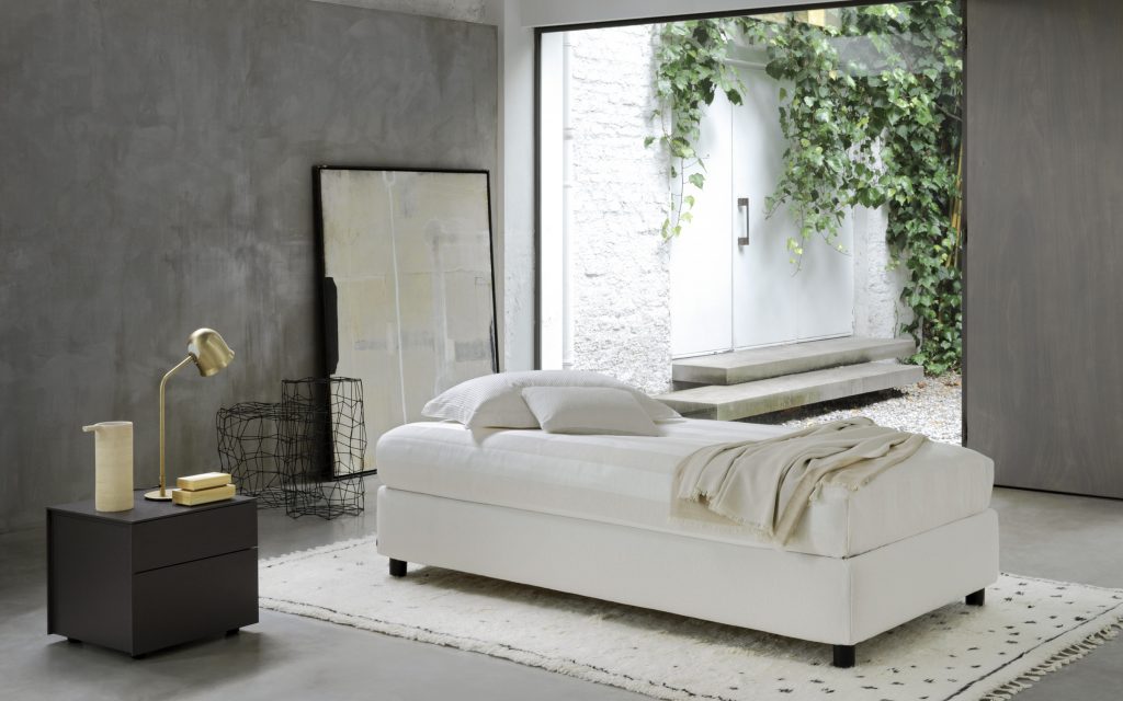 Sommier Single bed. White base with four black wheels and a mattress in a bed room.