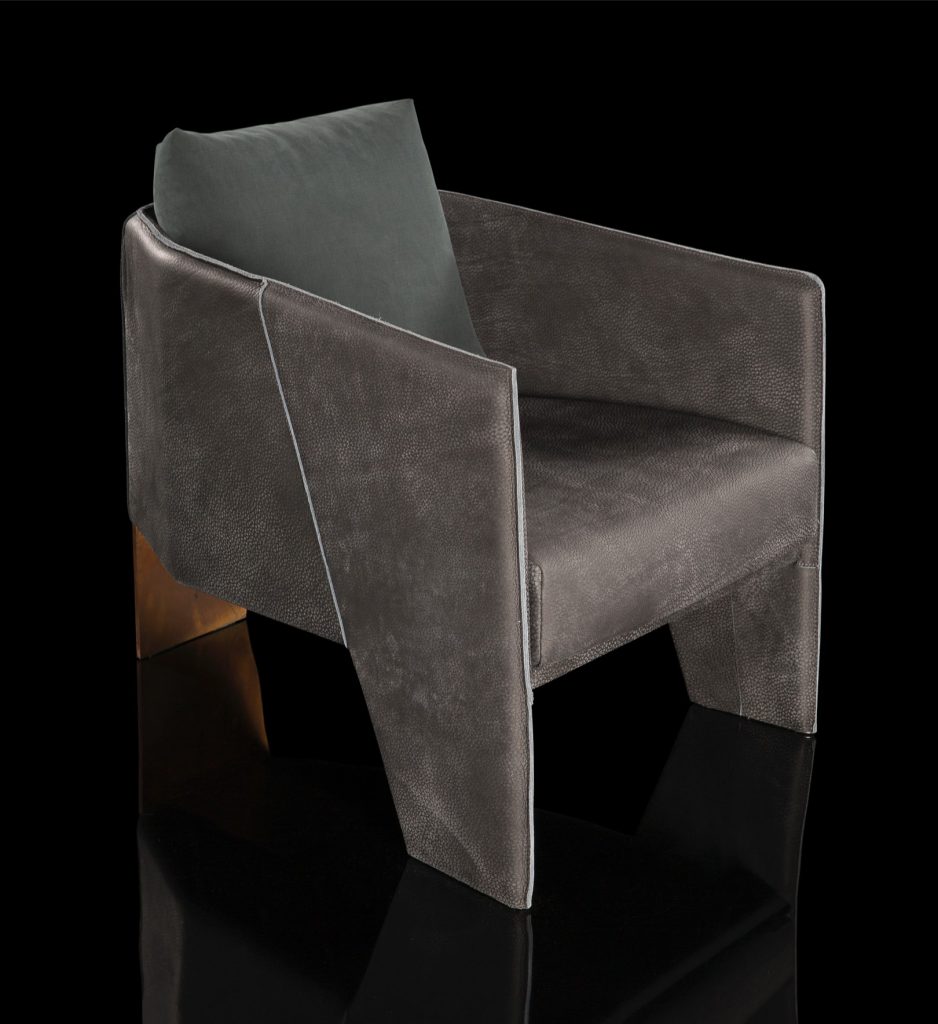 Ketch Armchair. Back leg in brass finish. finish padded seat, back, two legs and arms in gray leather finish, back cushion in gray fabric on a black background.