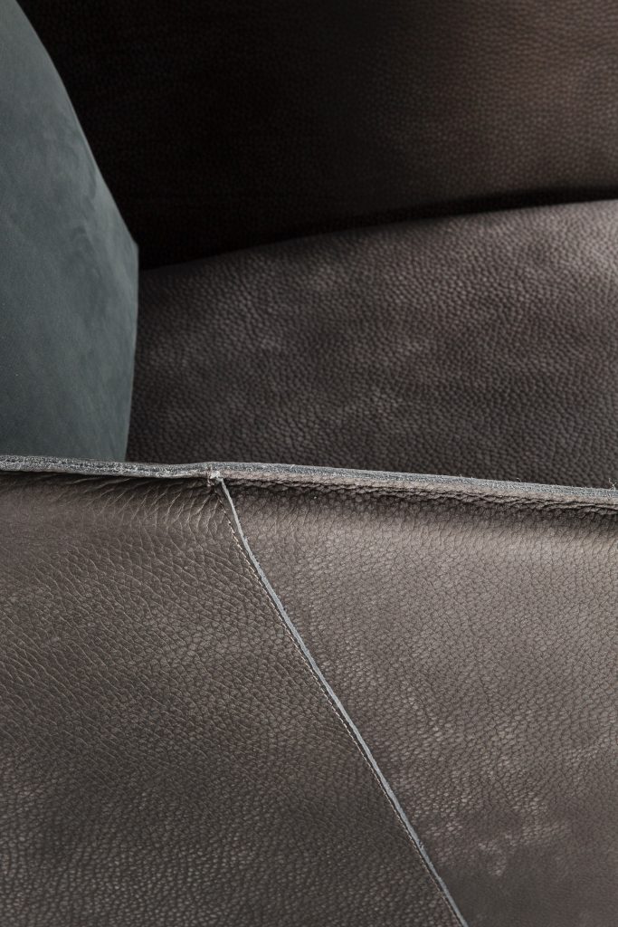 Closeup of Ketch Armchair. Back leg in brass finish. finish padded seat, back, two legs and arms in gray leather finish, back cushion in gray fabric on a black background.