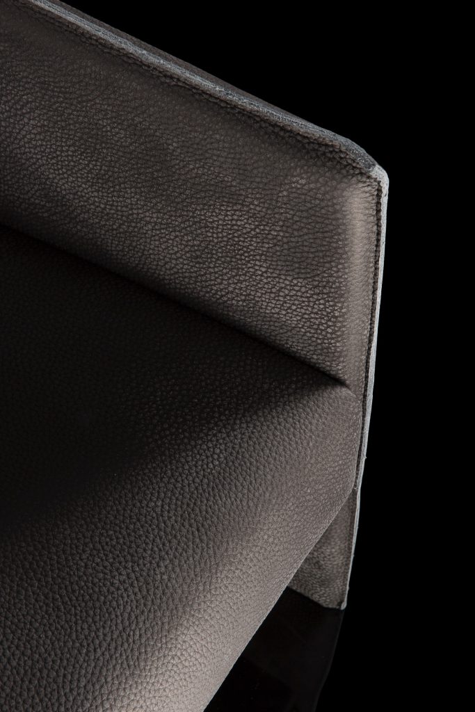 Closeup of Ketch Armchair. Back leg in brass finish. finish padded seat, back, two legs and arms in gray leather finish, back cushion in gray fabric on a black background.
