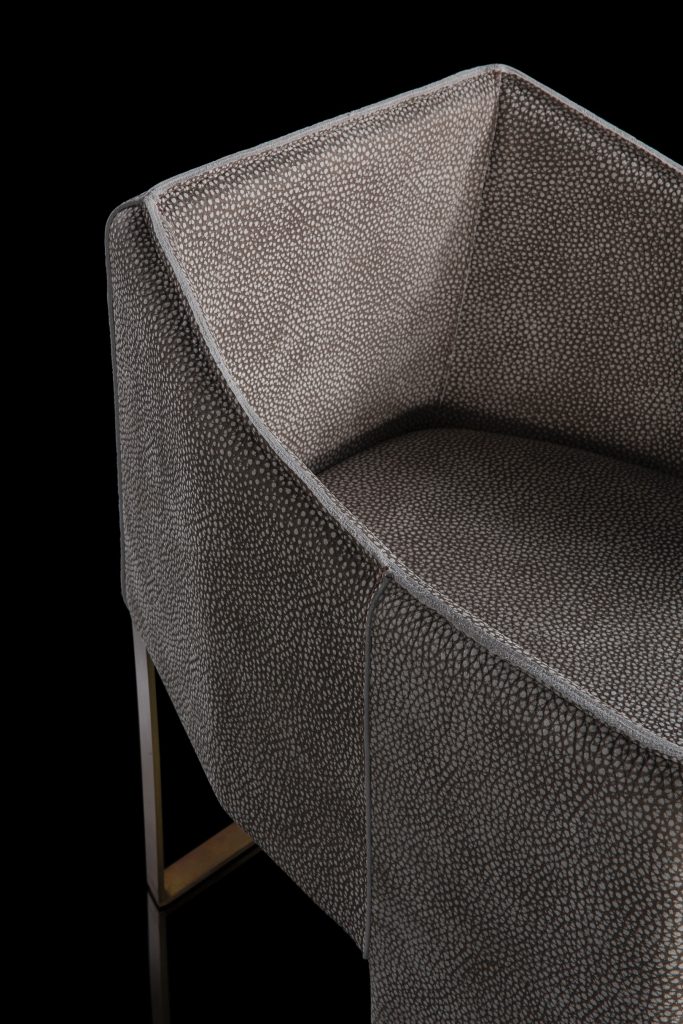 Closeup of Ketch Chair back leg in brass finish. finish padded seat, back, two legs and arms in gray leather finish, back cushion in gray fabric on a black background.