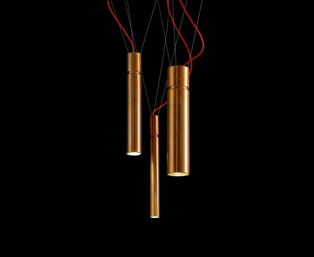 Three Ceiling Tubular Lights with a cylindrical shape in bronze suspended with ropes and a power cable covered in a red fabric on a black background.