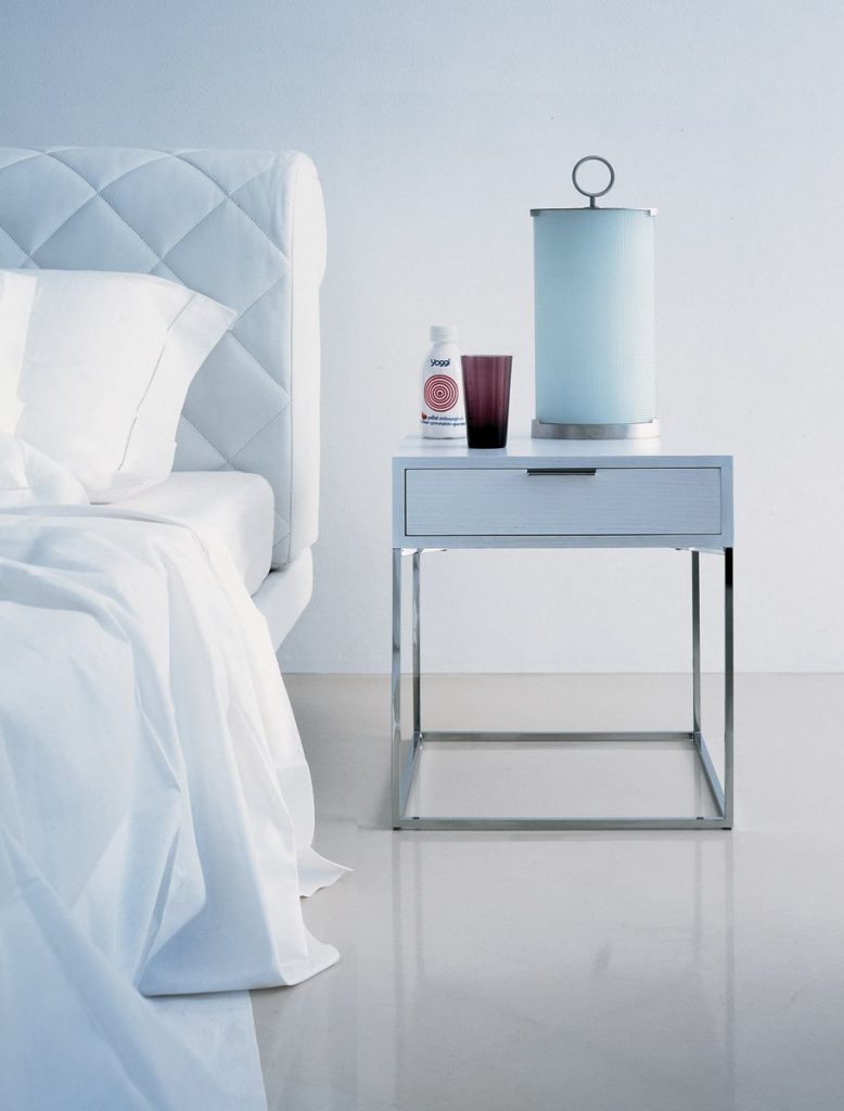 Oscar side table with a sleek silver frame and a white wooden top with drawer next to a bed.