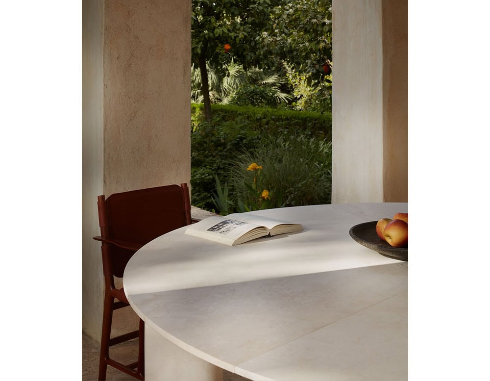 A Giotto Outdoor Dining Table with a round white top is partially visible with a chair and a view of greenery through an arched opening