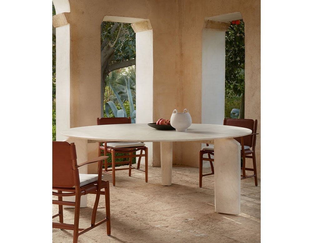 A Giotto Outdoor Dining Table with an oval white top and a central pedestal base is surrounded by chairs on a patio with arched openings