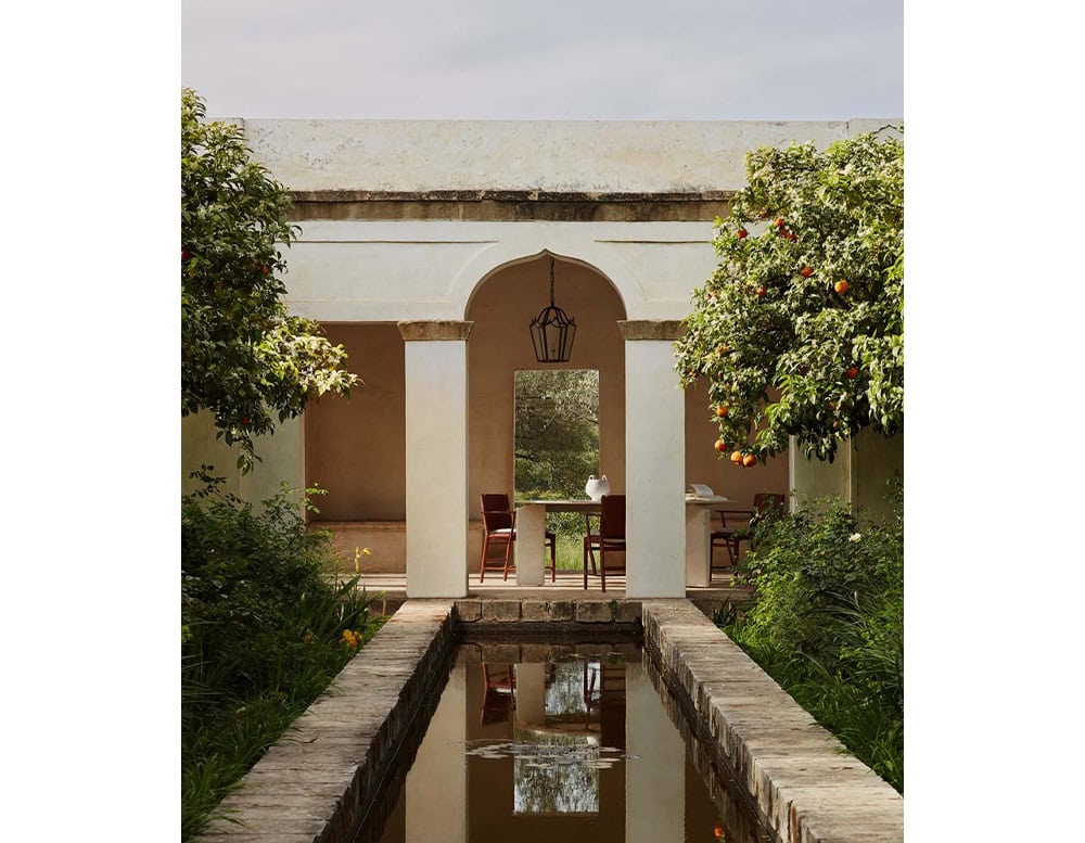 A Giotto Outdoor Dining Table is visible through an arched doorway in a courtyard with a reflecting pool and greenery