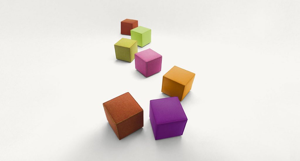 Seven Cubo pouf, one in purple, one in pink, one in brown, one in red, one in orange, one in green and one in yellow on a white background.