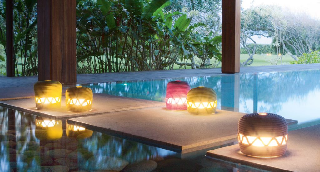 Four led Agadir lanterns made of rope cord in a spiral-like pattern, three in yellow, one in pink and one in white on a platform in a pool.