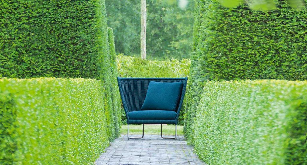 Blue Ami Outdoor Armchair, structure upholstery in fabric, seat cushion in fabric, structure and two legs in steel in a garden.