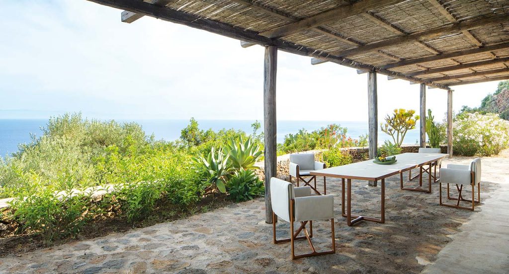 Four Portofino Outdoor Chairs with armrests and little armchair, structure and chair in natural heartwood, upholstered in beige fabric in a dining room.