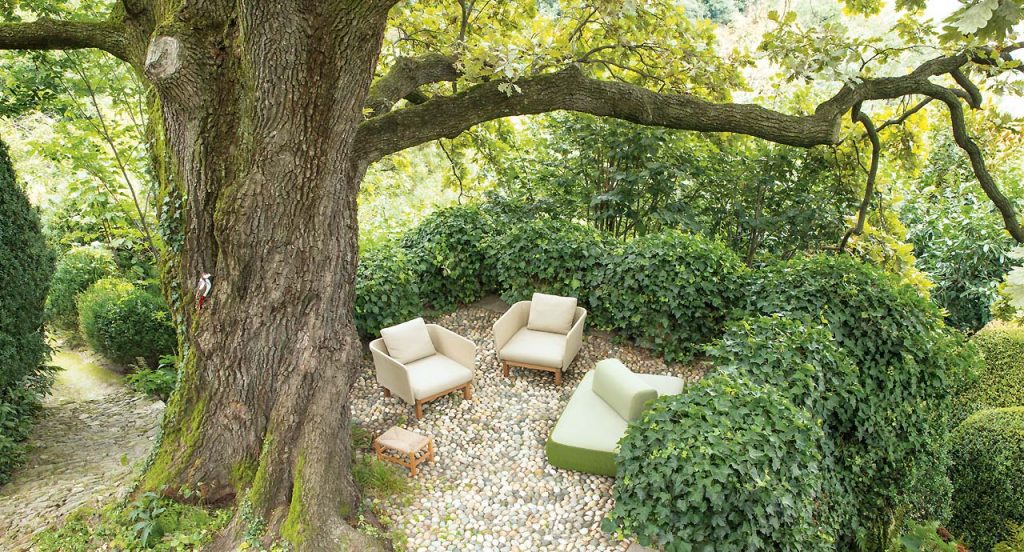 Two beige Sabi Armchairs, base and two legs made of natural wood, backrest in woven rope cord and cushion in fabric in a garden.