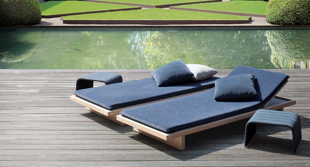 Two Sabi Sun Beds, structure in natural wood, seat pad in blue polyester next to a lake.