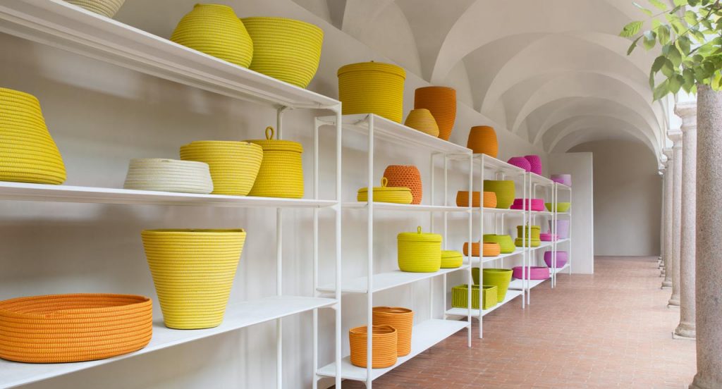 Sika baskets, vases, bowls and planter covers with different form and dimension made in Rope cord, seven in green, eight in orange, one in white, nine in yellow, six in pink and three in purple in a hallway.