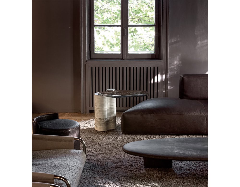 The Plynto coffee table stands by a window, featuring a white marble base and dark sculpted top. It complements a cozy living space with textured rugs, low seating, and warm natural light filtering through the trees.