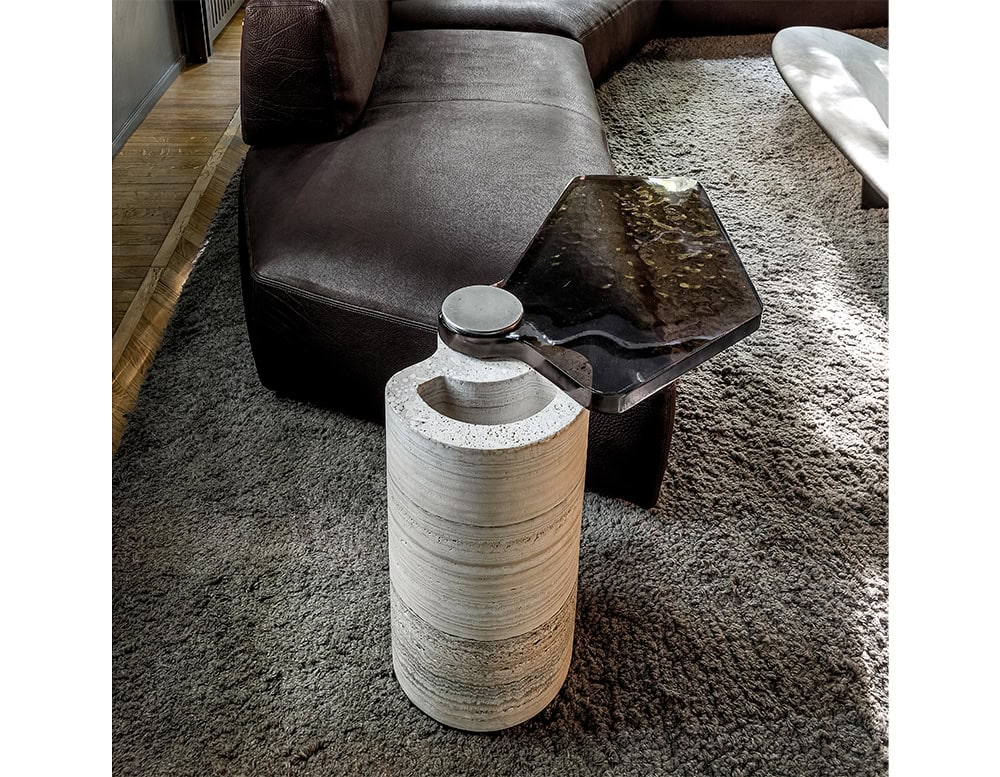 The Plynto coffee table pairs a cylindrical stone base with a dark, textured metal top, positioned beside a leather sofa in a refined living space with soft lighting and layered natural tones.