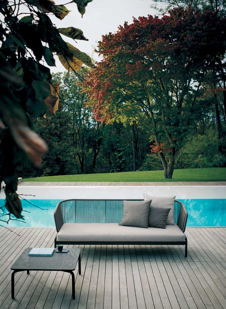 Spool Three Sofa in front of a pool on a grey colored deck with some fall trees in the background