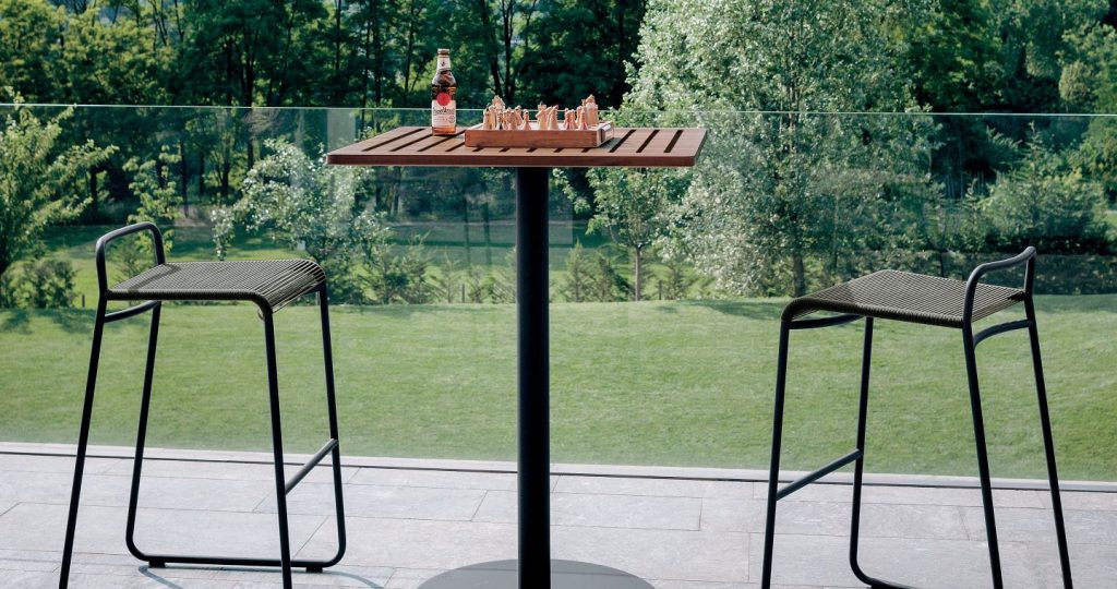Stem Eleven Bar Table with two barstools on either side of the table on a patio with grass and trees in the background