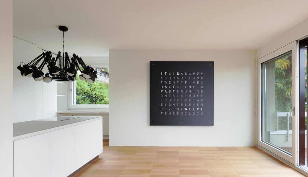QLOCKTWO One Hundred Eighty in black hanging on a white wall with a light wood floor and an island in front