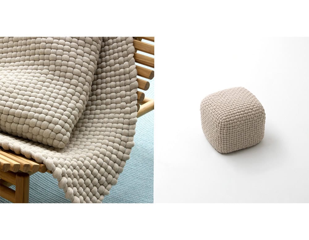 Beige ottoman with a cube shape and textured woven surface, placed beside a matching thick woven fabric draped over a wooden bench.