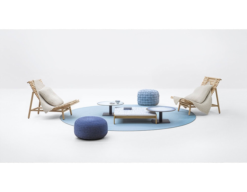 Minimalist lounge setup with two wooden chairs draped in knitted throws, blue ottomans, and low round tables arranged on a light blue rug.