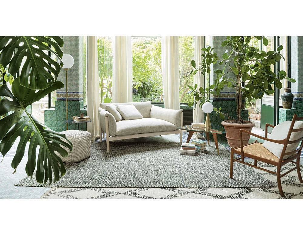 Inviting lounge area featuring a cream sofa, woven ottoman, and greenery-filled decor, with natural light streaming through tall windows onto a patterned rug.