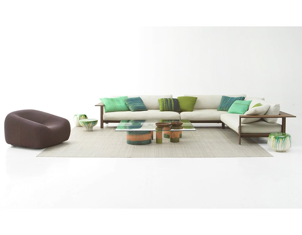 A spacious outdoor sofa with a wooden frame and cream cushions, accented by green and turquoise pillows, sits alongside sculptural tables and decor, creating a fresh, relaxed modern setting.