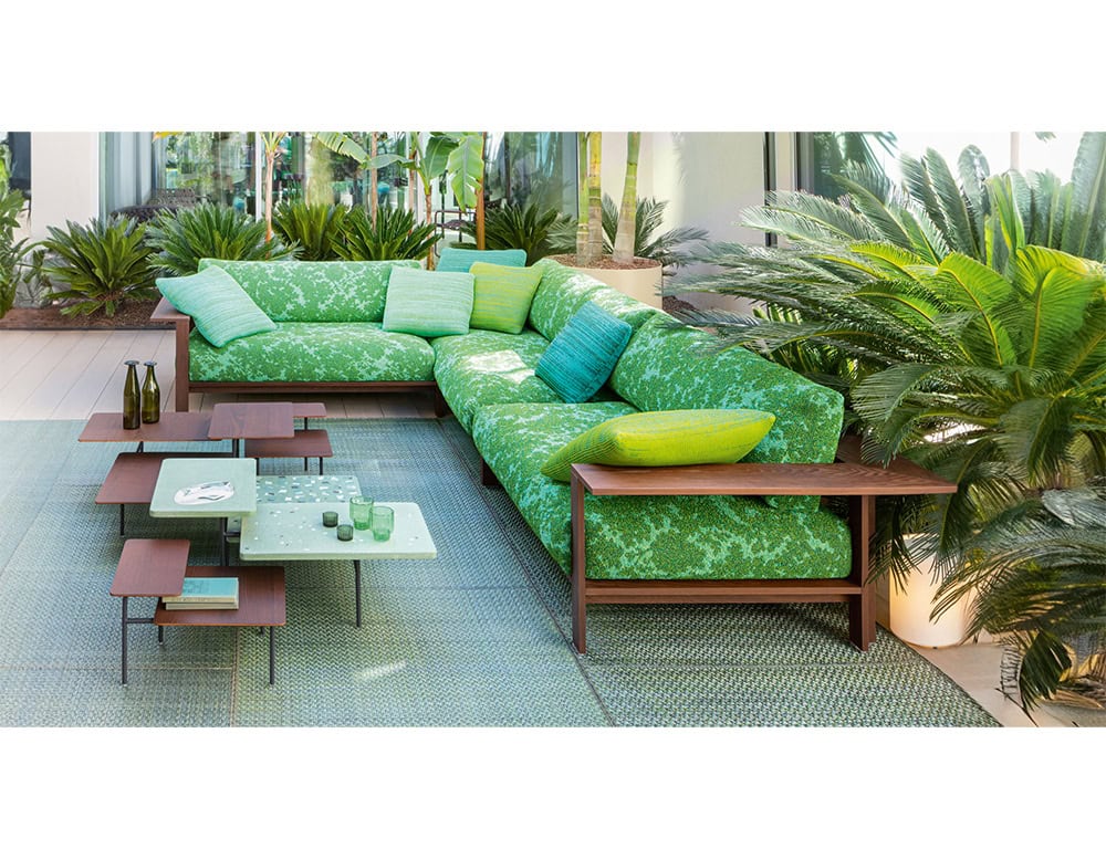 A vibrant outdoor lounge with an L-shaped green patterned sofa and wooden frame, paired with colorful cushions and modern low tables, surrounded by lush tropical plants and soft natural light.