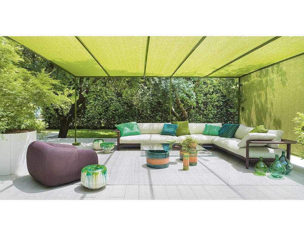 An inviting outdoor lounge with a cream L shaped sofa and green cushions under a light filtering canopy, surrounded by lush trees, colorful decor, and glass accents that enhance the serene garden setting.