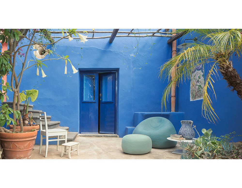 Softly rounded poufs in cool tones rest against vivid blue walls, offering a calming contrast. Their sculptural forms create an inviting corner for relaxation in this colorful courtyard.