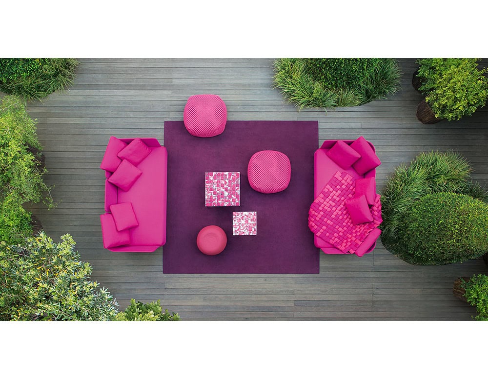 Modern outdoor living space featuring magenta sofas, matching cushions, and poufs on a rich purple base, balanced by natural green surroundings.