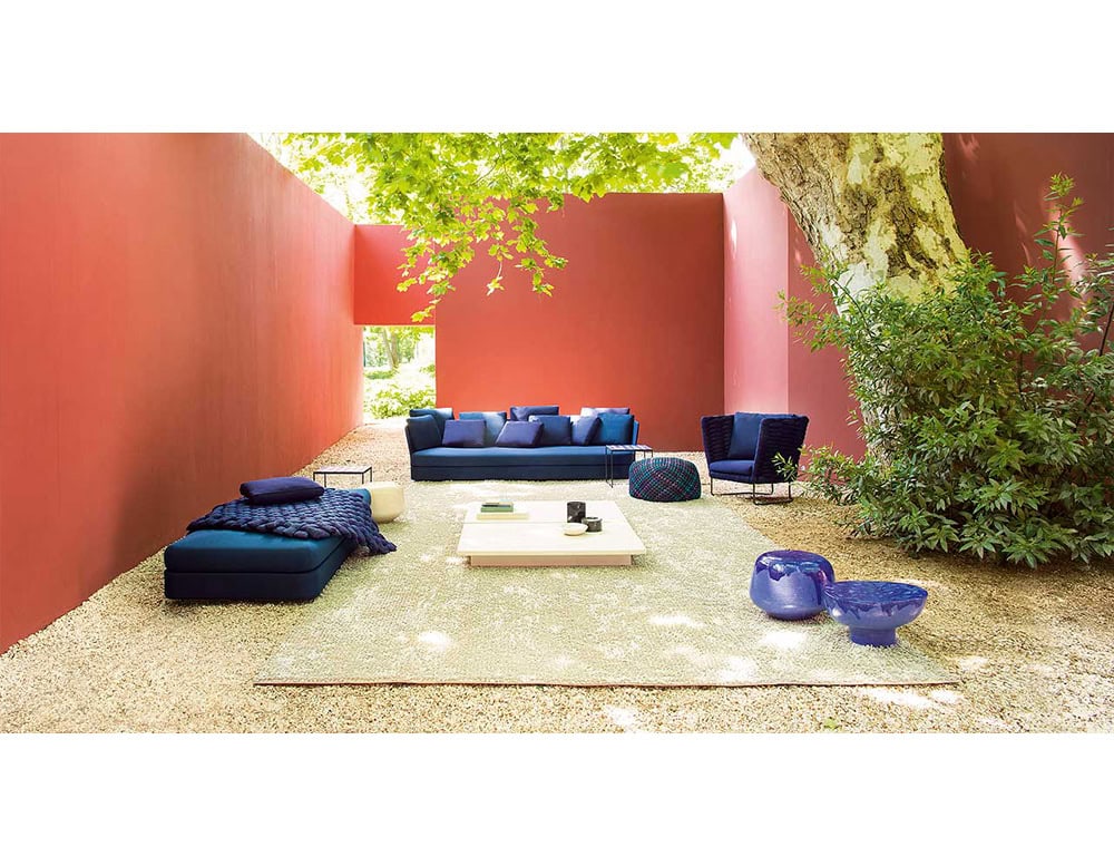 Modern outdoor lounge featuring deep blue sofas and accessories, framed by terracotta walls and shaded by lush trees for a vibrant yet inviting retreat.