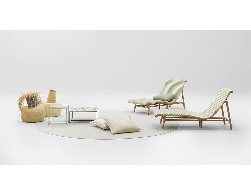 Wooden loungers with slatted design and soft cushions are paired with low tables, woven baskets, and floor pillows over a round rug for a balanced composition.