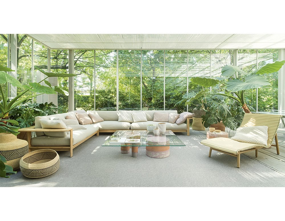 A neutral sectional sofa and wooden lounge chair with pillows anchor the space, complemented by baskets and a glass coffee table with ceramics, framed by floor to ceiling windows that open to a green view.
