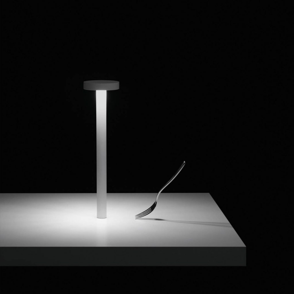 Tetatet table lamp in front of a black background illuminating a white table with a glass of water next to it