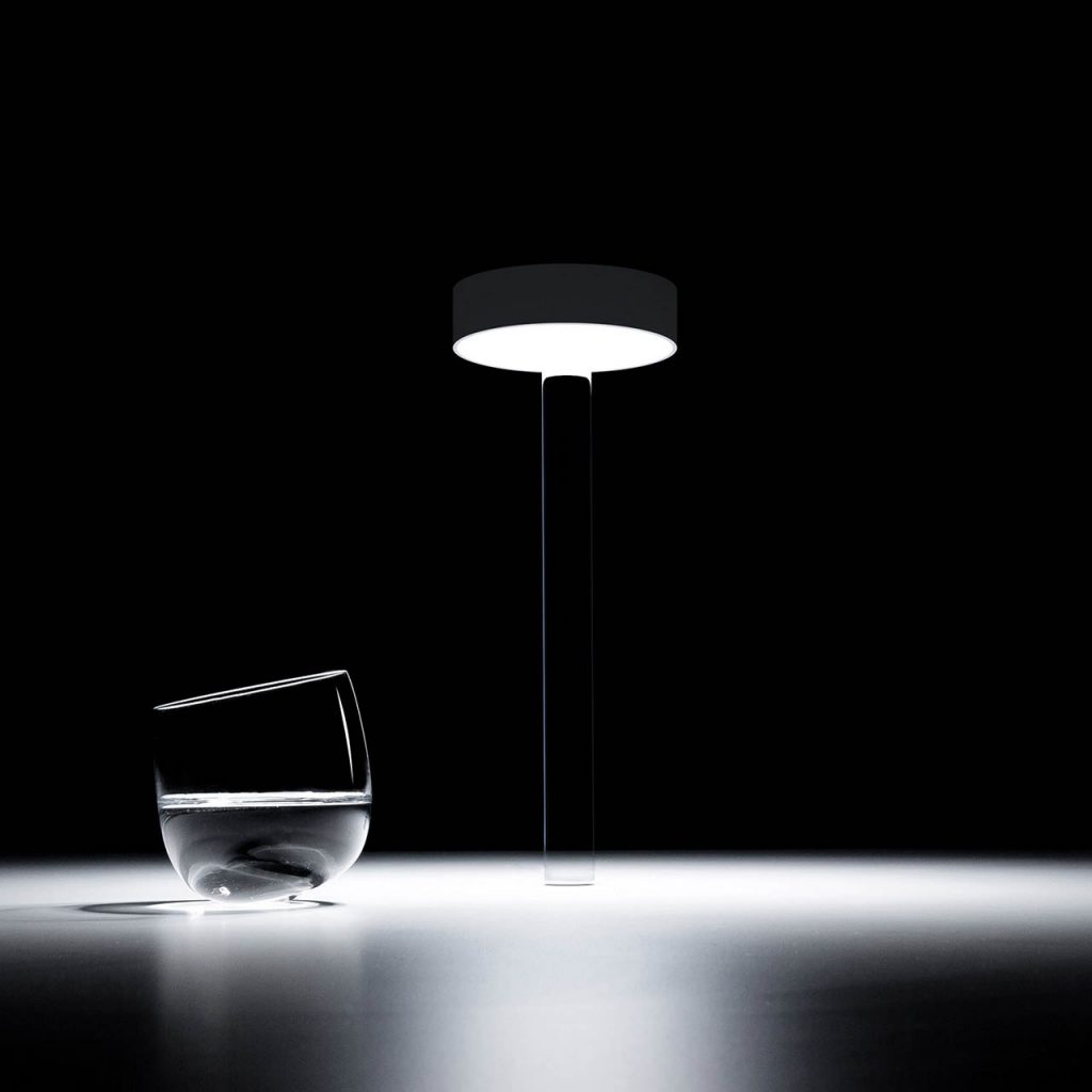 Tetatet flute light in front of a black background illuminating a white table with a glass of water next to it