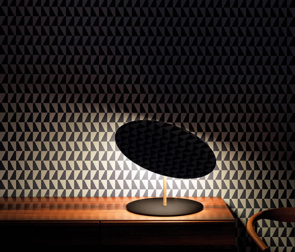Calvino lamp illuminating a wooden table with a funky patterned wall in a dark room
