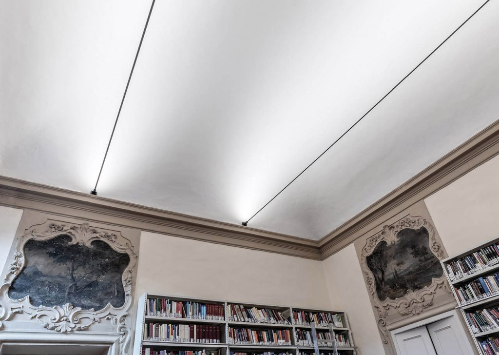 Two Infinito lights parallel to each other illuminating a white ceiling with bookshelves underneath