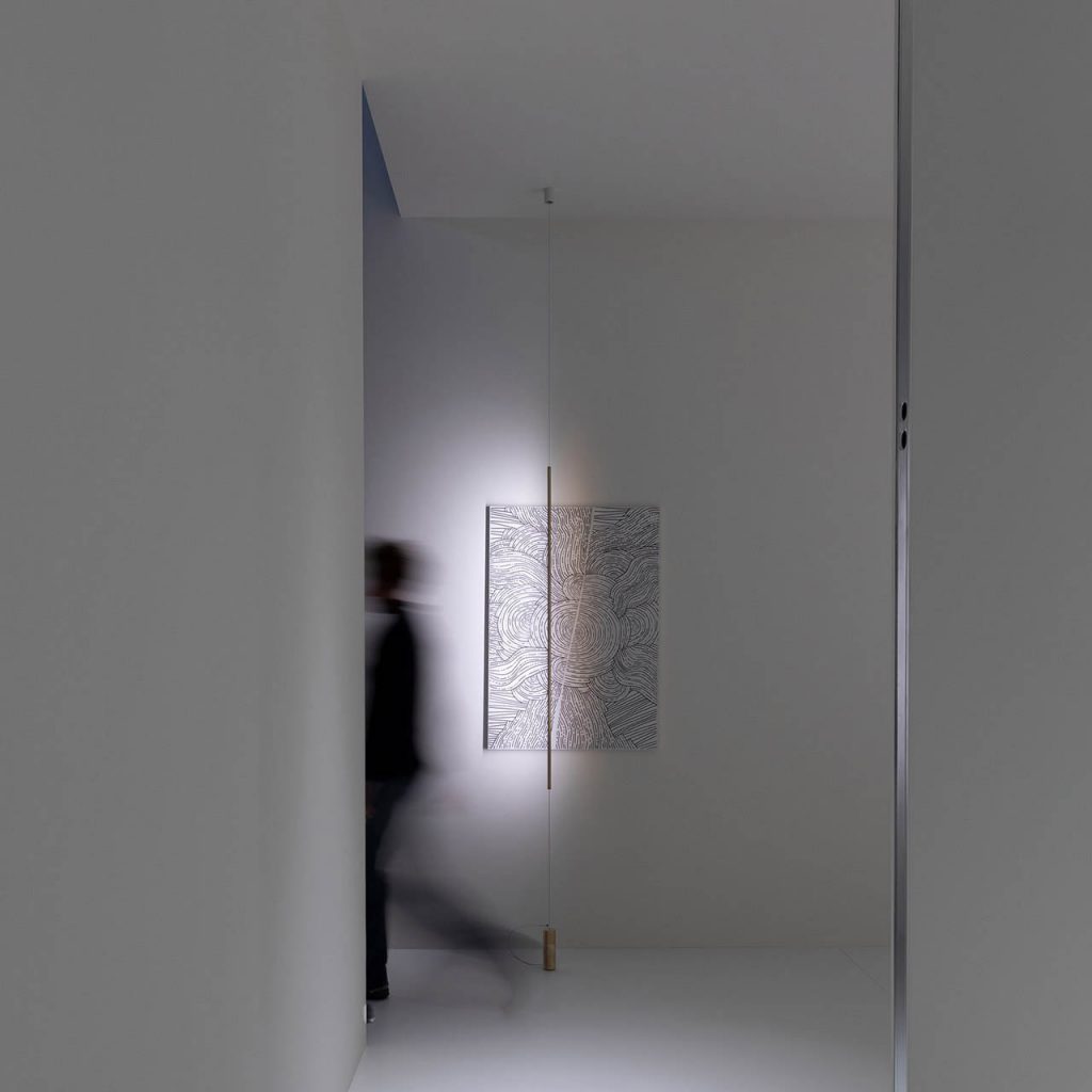 Masai light shining on an art work at the end of the room on a white wall with a person walking in front