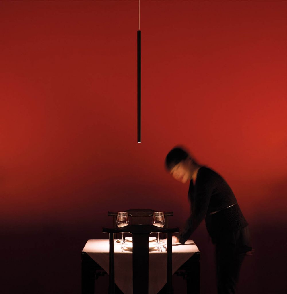 Miss suspension lamp hanging over a small table in front of a red wall with a person sitting at the table