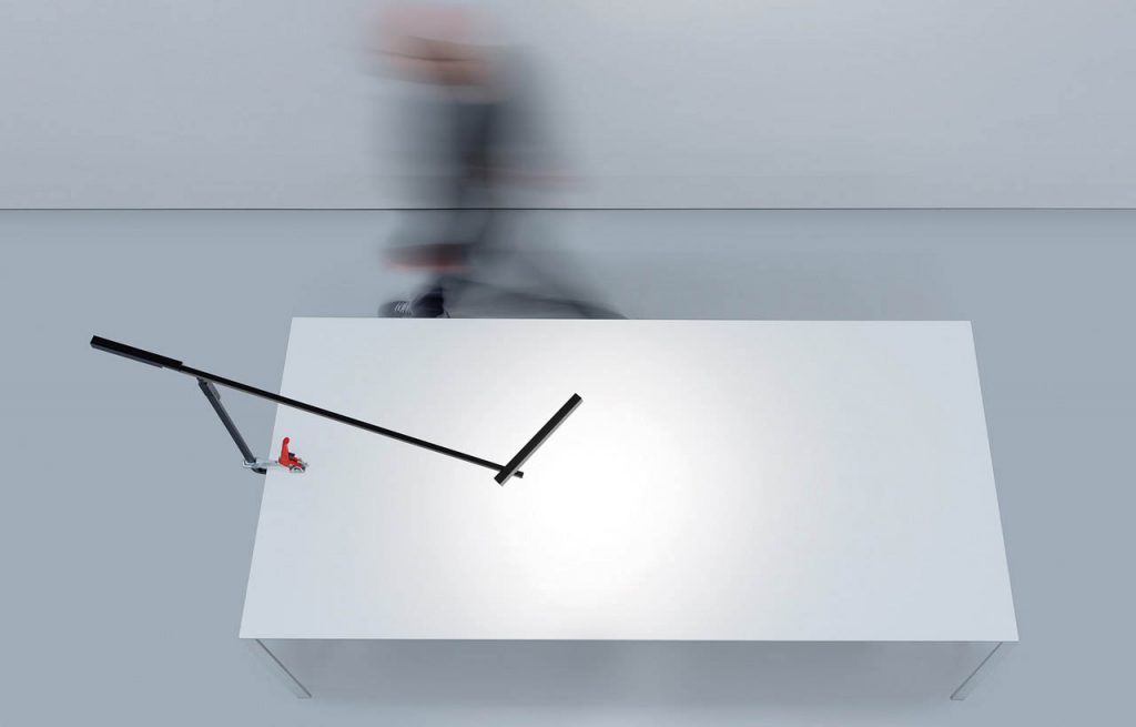 Morsetto desk lamp illuminating a white desk with a person walking behind the table