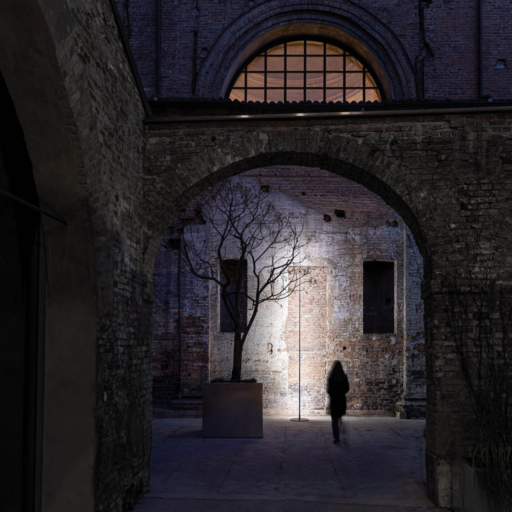 Origine Outdoor shining against a grey wall with a person standing behind the light and an archway in the foreground