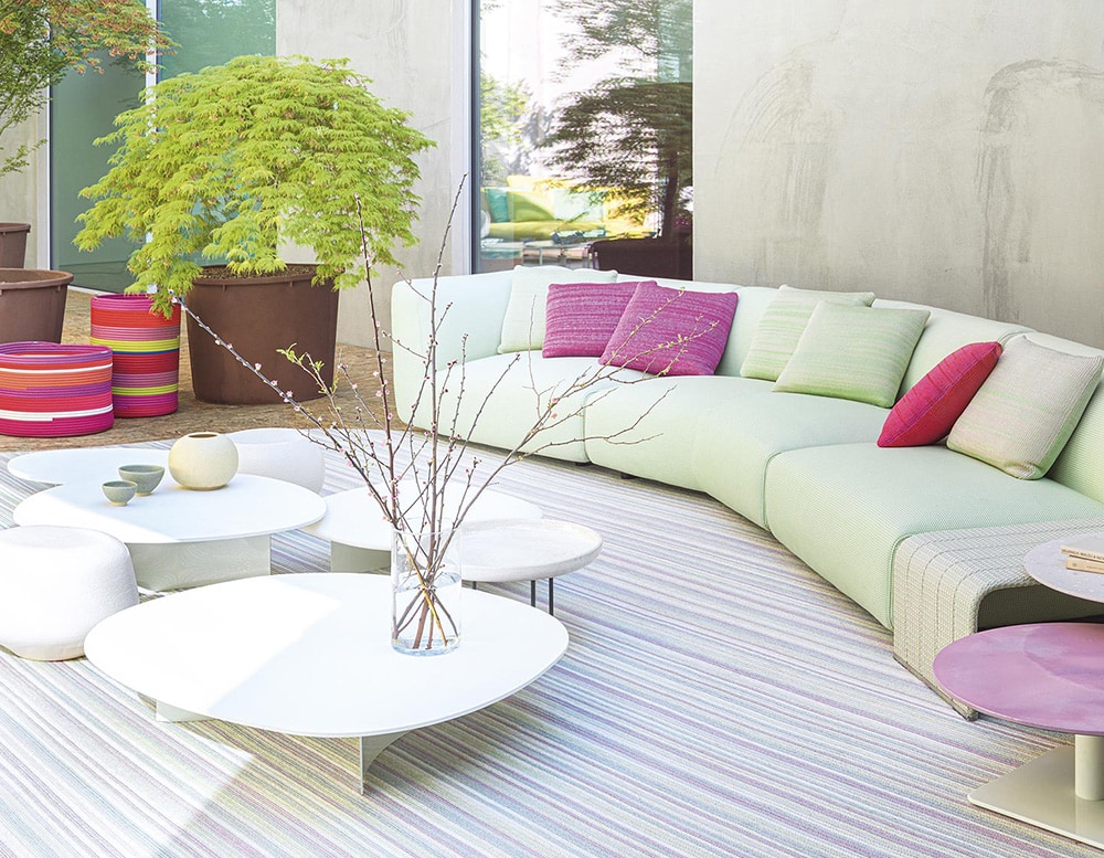 Isole table in front of a green sofa on top of a stripped rug with windows in the background